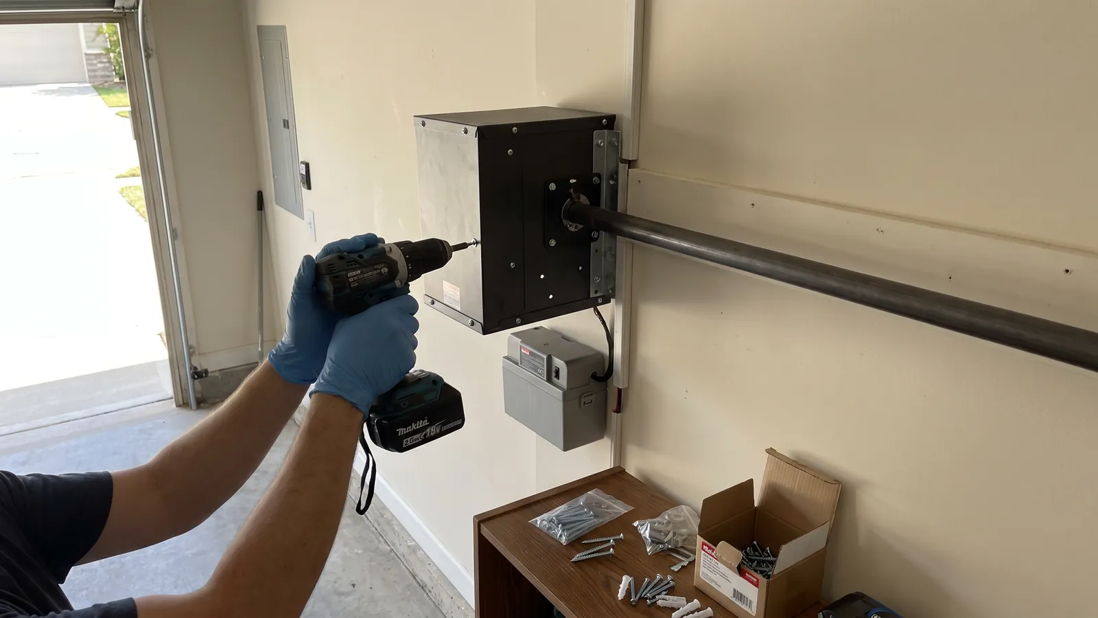 Technician mounting a jackshaft wall-mount garage door opener with a battery backup beside the torsion shaft in a finished two-car garage